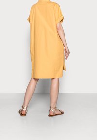 Abito di cotone giallo a maniche corte con spacchi laterali, taglio dritto e colletto. Indossato con sandali pianeggianti marroni, che lasciano scoperti i piedi.