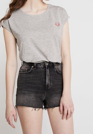 Woman wearing a light grey t-shirt with a small red crab logo and high-waisted black denim shorts with frayed hems.