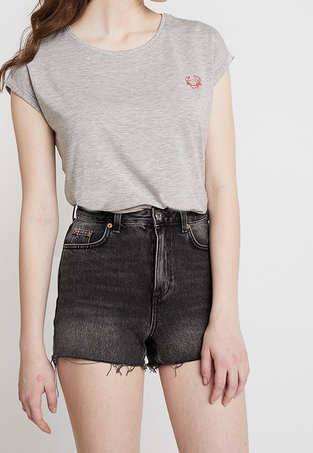 Femme portant un t-shirt gris clair avec un petit logo crabe rouge et un short en jean noir taille haute avec des bords effilochés.