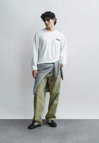 Άσπρο μακρυμάνικο πουκάμισο με λογότυπο, γκρι πουλόβερ δεμένο στη μέση, χακί φαρδιά παντελόνια και μαύρα αθλητικά παπούτσια με λευκές ρίγες. Μαύρα γυαλιά ηλίου.