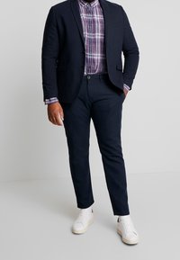 Námořnický blazer přes kostkovanou košili v purpurové a bílé barvě, doplněný námořnickými kalhotami a bílými teniskami. Hladká textura, upravený střih a jemné detaily.