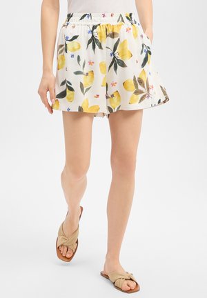 Femme portant un short blanc à imprimé floral jaune et des sandales beiges, marchant avec les mains dans les poches devant un fond uni.