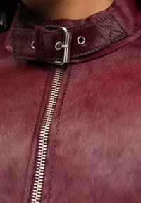 Veste en cuir bordeaux avec une texture lisse, dotée d'une fermeture éclair en argent et d'une fermeture à boucle au col pour un détail supplémentaire.