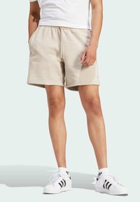 Beige bomullsshorts med elastisk midja, sidofickor och rak skärning, tillsammans med vita sneakers med svarta ränder.