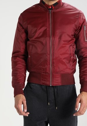 Homme portant une veste bomber rouge zippée avec des poignets et un bas côtelés, associée à un pantalon noir à cordon.