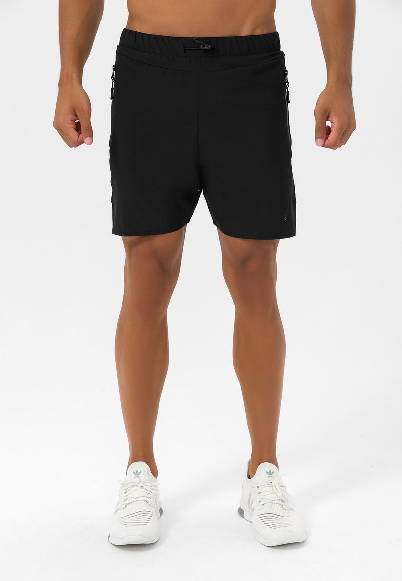 Zwarte sportshorts met een elastische tailleband, ritszakken aan de zijkant en een strakke pasvorm. Gecombineerd met witte sneakers.