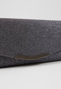 Bolso de mano en tela gris oscuro con un acabado texturizado; cuenta con un acento metálico curvado en el cierre.