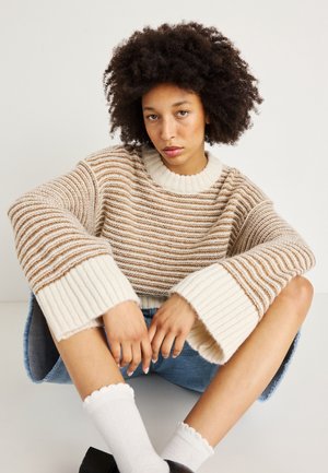 Gestreifter Pullover in Creme und Braun, mit einem gerippten, oversized Kragen und Bündchen. Das Modell kombiniert ihn mit einem Jeansrock und weißen Socken.