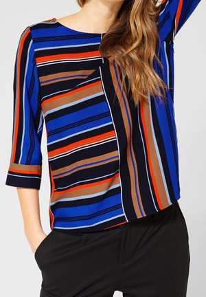Kobieta ubrana w długi rękaw w paski w kolorach niebieskim, czarnym, brązowym, pomarańczowym i jasnoniebieskim, w czarnych spodniach, z jedną ręką w kieszeni, na jednolitym tle.