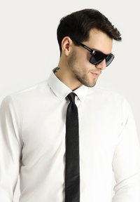 Gafas de sol negras, con montura rectangular, combinadas con una camisa blanca y una corbata negra con un patrón sutil. Textura suave y diseño elegante.
