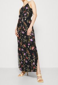 Robe maxi en tissu noir avec un motif floral comportant des fleurs roses, violettes et blanches. Comprend une taille ceinturée et une fente sur le côté.