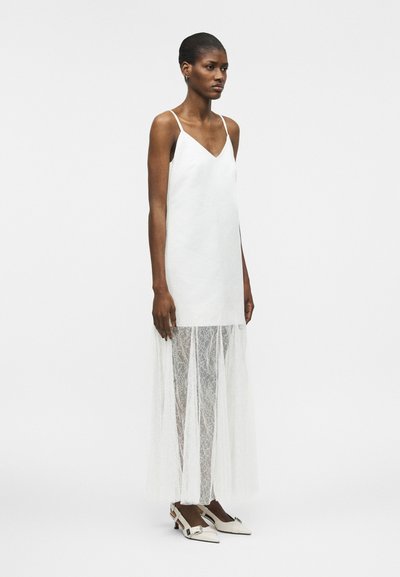 Γυναίκα με άσπρο αμάνικο φόρεμα με δαντέλα που εκτείνεται από το μέσο του μηρού μέχρι το πάτωμα και άσπρα παπούτσια με χαμηλό τακούνι σε απλό φόντο.