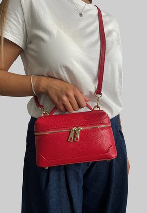 Borsa a mano in pelle rossa con finitura testurizzata, accessori in oro, chiusura a doppia zip e tracolla regolabile, tenuta in mano su uno sfondo semplice.