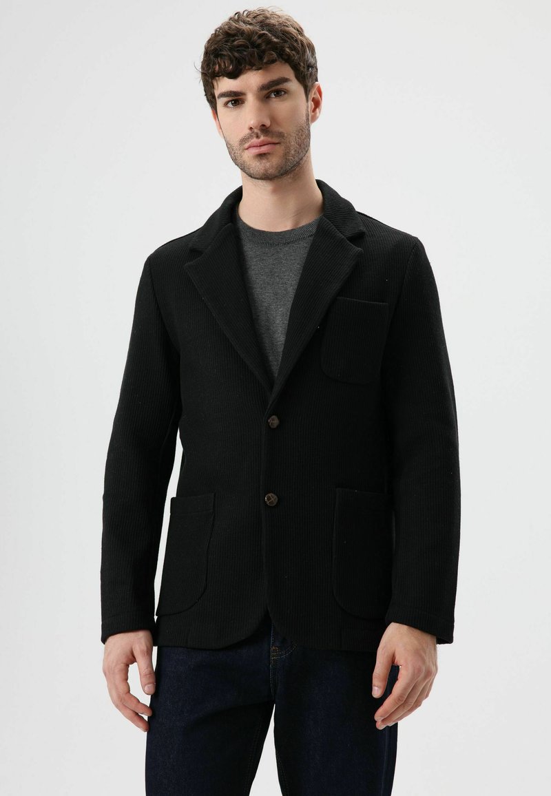 Zwarte ribgebreide blazer met een kraag met inkeping, twee steekzakken aan de voorkant en bruine knopen. Draagt over een grijs overhemd. Getailleerd ontwerp, gestructureerde stof.