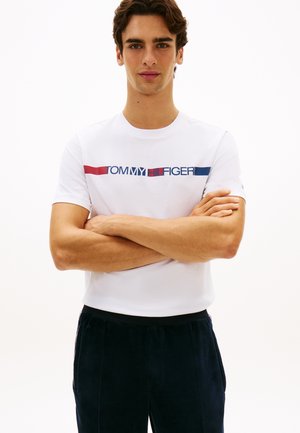 Muškarac sa sklopljenim rukama, nosi bijelu Tommy Hilfiger majicu s crvenim, bijelim i plavim logom i crne hlače na jednostavnoj bijeloj pozadini.