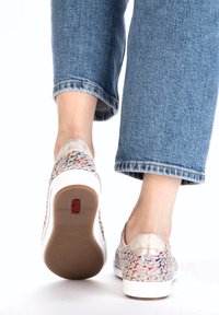 Baskets tissées multicolores avec un accent métallique au talon, semelle blanche et surface texturée, assorties à un jean en denim bleu retroussé.