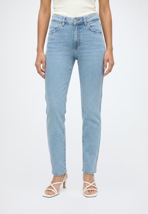 Personne portant un jean skinny taille haute bleu clair et des sandales blanches à talons avec lanières, debout devant un fond blanc uni.