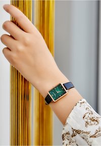 Rektangulär klocka med gr�ön urtavla, roséguldiga detaljer och romerska siffror på ett svart läderarmband, bärs på en hand mot gyllene stänger.