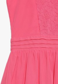 Close-up van een roze jurk met een kant bodice, drie horizontale plooien bij de taille en een gathered rok detail.