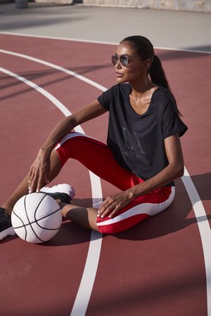 Frau mit Sonnenbrille sitzt auf einem Freiluft-Basketballplatz, trägt schwarzen Top und rote Leggings, legt die Hand auf einen weißen Basketball.