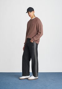 Ruskea texturoitu neule, jossa on ribbireunukset hihoissa, yhdistetty tummiin farkkuihin, joissa on sivupinnat ja valkoiset sneakrit. Musta lippis päässä.