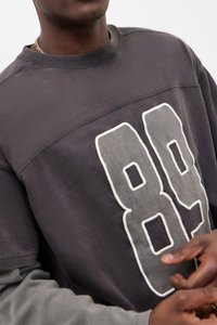 Γκρίζα ζακέτα με μεγάλο λευκό "89" εκτυπωμένο στο μπροστινό μέρος, με στρογγυλή λαιμόκοψη και υφή υφάσματος. Μακριές γκρίζες μανσέτες από κάτω.