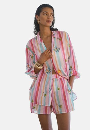 Frau trägt ein pastellfarben gestreiftes Hemd- und Shorts-Set mit Sonnen- und Palmenmotiven, dazu goldene Armreifen und sonnenförmige Ohrringe.