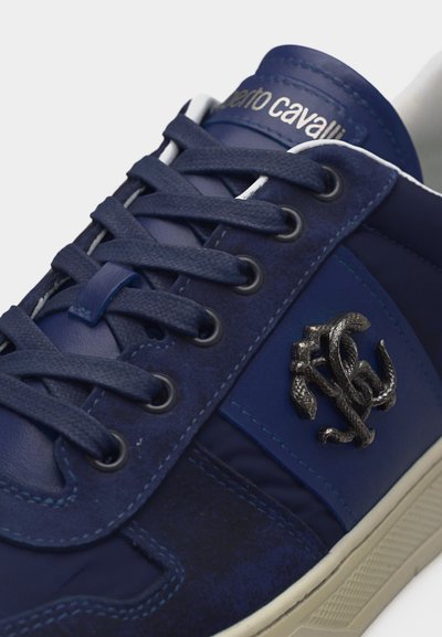 Σκούρο μπλε αθλητικό παπούτσι Roberto Cavalli με ανάγλυφο μεταλλικό λογότυπο, χοντρές κορδόνια και εκρού ελαστική σόλα, κοντινή λήψη στο επάνω μέρος και τη γλώσσα.