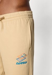 Bežiniai sportiniai kelnaičiai su elastinga juosmens juosta, šoninėmis kišenėmis ir mėlynu bei oranžiniu logotipu kairėje kojoje. Minkšta, tekstūruota medvilnė.