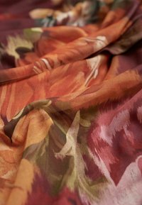 Gelaagd textiel met een bloemmotief in warme tinten van oranje, rood en groen, gedrapeerd om het vloeiende ontwerp te tonen.