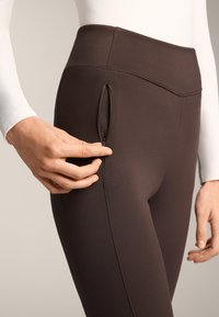 Bruine leggings met een hoge taille en een zijritszak. Glad, rekbaar stof met subtiele naden. Hand die naar de rits reikt.