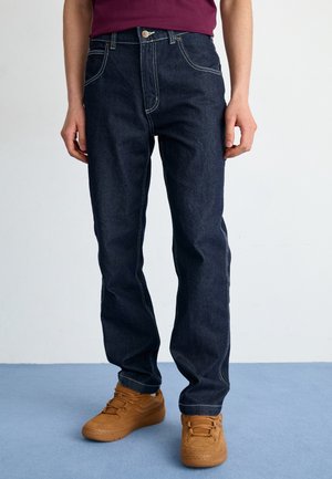 Person iført mørkeblå jeans med hvide syninger og lysebrune sneakers står på en blå gulv.