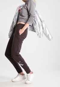 Felpa grigia corta, pantaloni della tuta neri con logo bianco e sneakers bianche. Giacca bomber argentata appoggiata sulle spalle. Texture lisce.