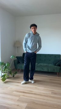 Jauns cilvēks, tērpies gaiši pelēkā kapučjakā un tumšās biksēs, stāv uz koka grīdas blakus zaļai dīvānam un istabas augam.