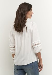 Camicia bianca con maniche a tre quarti voluminose, dettagli in pizzo intricato sulle maniche e vestibilità rilassata, abbinata a jeans blu.