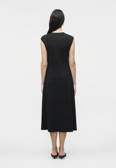 Γυναίκα με μακριά σκούρα μαλλιά, που φοράει μαύρο μίντι φόρεμα χωρίς μανίκια και μαύρα σανδάλια με χαμηλό τακούνι, στέκεται με την πλάτη προς τα εμπρός σε λευκό απλό φόντο.