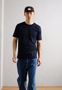 Tricou din bumbac de culoare bleumarin, cu mâneci scurte și guler rotund, purtat cu blugi din denim de culoare albastru deschis și o șapcă maro cu un patch logo.