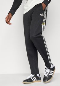 Μαύρο αθλητικό παντελόνι με λευκές ρίγες στο πλάι, με κεντημένο λογότυπο σε κίτρινο χρώμα. Συνδυασμένο με μαύρα και λευκά αθλητικά παπούτσια.