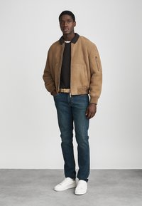 Barna velúrbombázó dzseki fekete gallérral, elülső zsebekkel és cipzáras ujjakkal. Fekete pulóver és kék farmer fölött viselve, fehér tornacipővel kiegészítve.