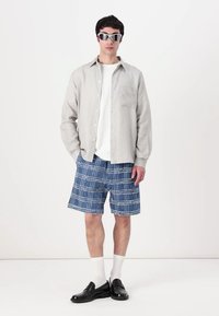 Grau kariertes Button-Up-Shirt über einem weißen T-Shirt, kombiniert mit blauen Karo-Shorts, weißen Socken und schwarzen Slippern. Silberne Sonnenbrillen runden den Look ab.