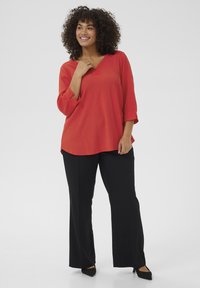 Blouse rouge à manches longues avec un décolleté en V, fentes latérales et une finition texturée, associée à un pantalon droit noir et des talons noirs.
