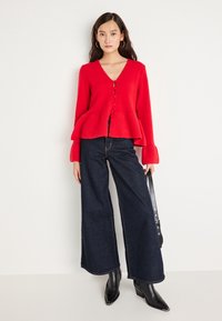 Cardigan tricotat roșu cu decolteu în V și tiv peplum, având nasturi pe față. Asortat cu blugi întunecați cu picioare largi și botine negre cu toc.