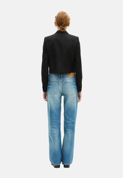 Femme avec les cheveux attachés, portant un blazer noir court et un jean taille haute bleu clair, se tenant de dos contre un fond blanc.