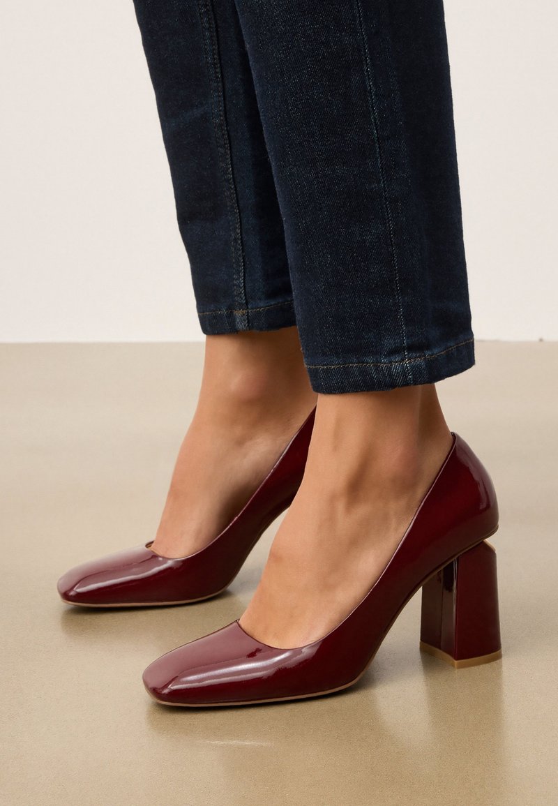 Burgundy lackläder högklackade skor med fyrkantig tå och klumpig klack, i kombination med mörk denim, liksnittade byxor.