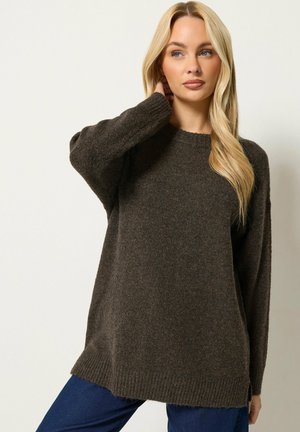 Lyshåret kvinde med langt hår iført en løs mørkebrun strikket sweater og blå bukser, poserer med den ene hånd nær halsen.