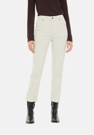 Persona que lleva pantalones rectos de tiro alto color blanco roto, camiseta oscura de manga larga y botas negras hasta el tobillo, de pie sobre fondo blanco.