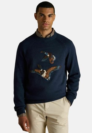Marineblauer Strickpullover mit einem strukturierten Muster aus fliegenden Vögeln in Braun und Creme. Gerippter Kragen und Bündchen, über ein kariertes Hemd getragen.