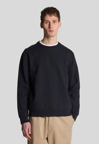 Felpa navy in un tessuto morbido e testurizzato, con collo a giro e maniche lunghe, completata da un orlo dritto. Indossata sopra pantaloni beige chiaro.