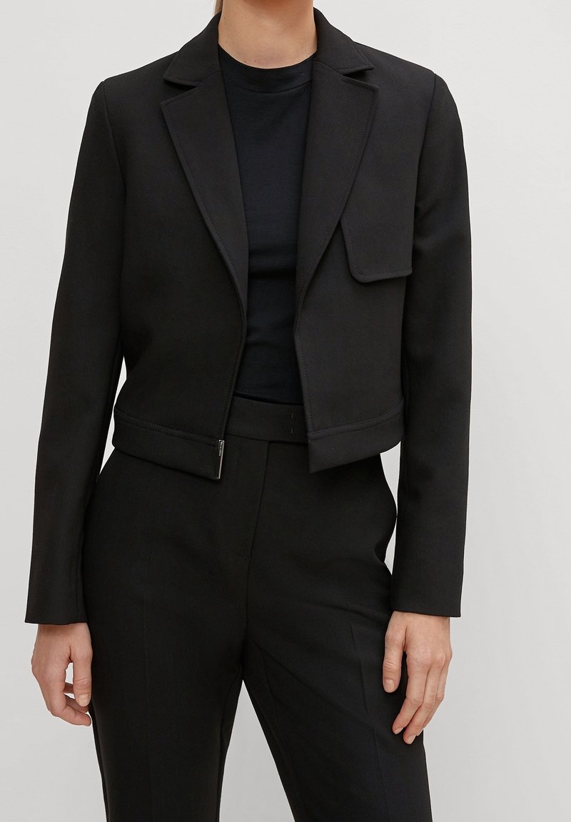 Musta lyhyt blazer, jossa on lovettu kaulus, etuläppäpussi ja sileä pinta, yhdistettynä mustaan crew neck -paitaan ja mittatilaustyönä tehtyihin housuihin.