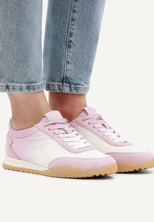 Person wearing light pink and white sneakers with gum soles paired with cuffed light blue jeans on a white background.